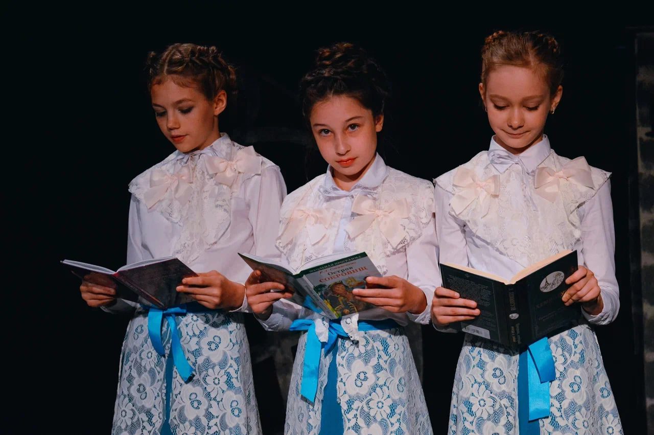 Три девочки в театральных платьях стоят на сцене и читают книги